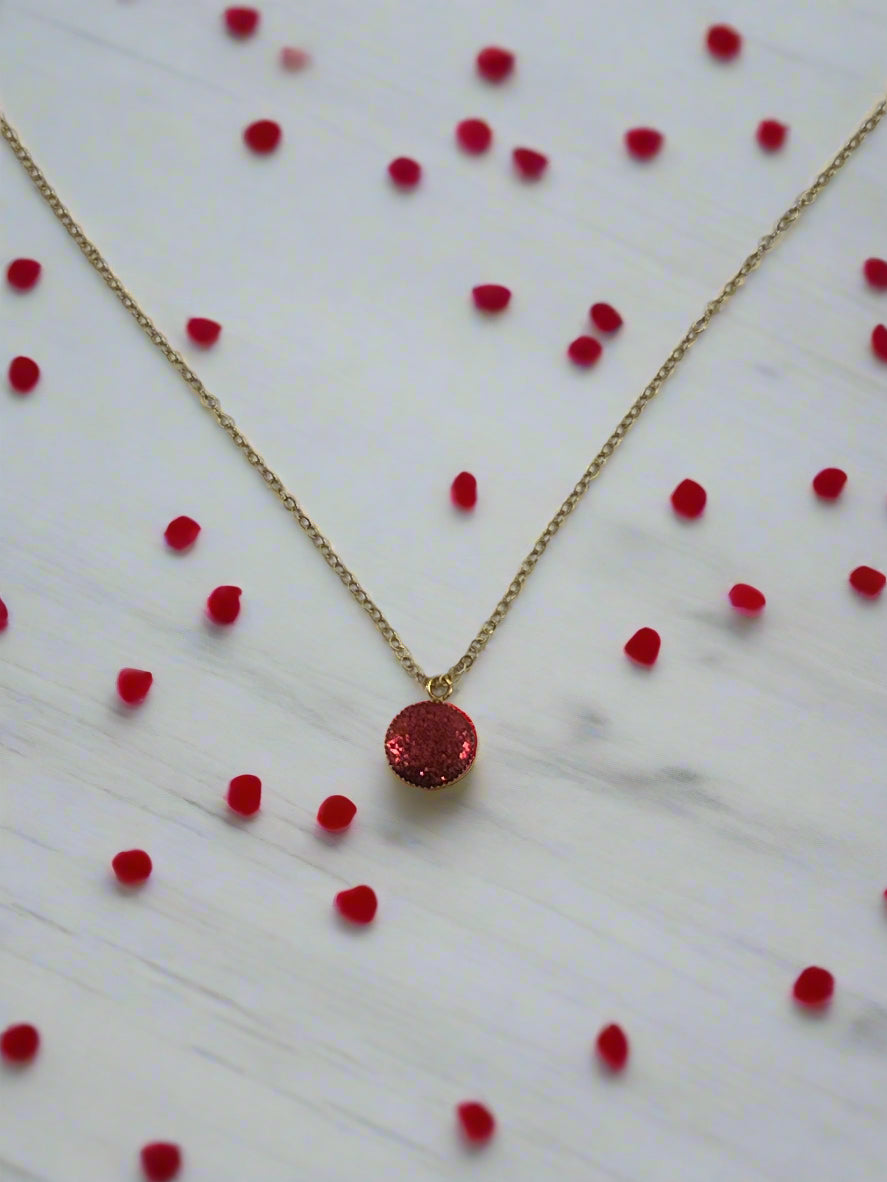 Ruby Crimson Shimmer Charm Necklace on a gold chain with red gems scattered on a marble surface.
