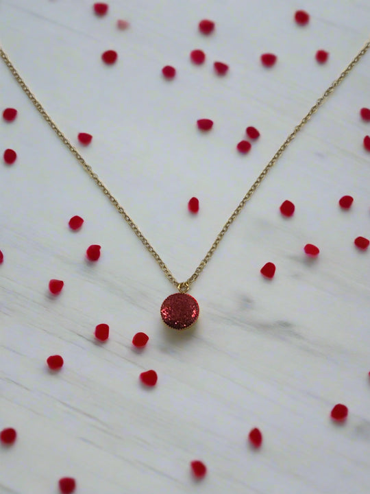 Ruby Crimson Shimmer Charm Necklace on a gold chain with red gems scattered on a marble surface.
