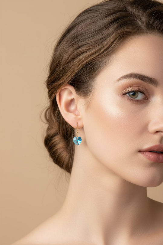 A pair of gold-tone dangle earrings with small, heart-shaped blue crystals, displayed on a light-colored wooden surface.
