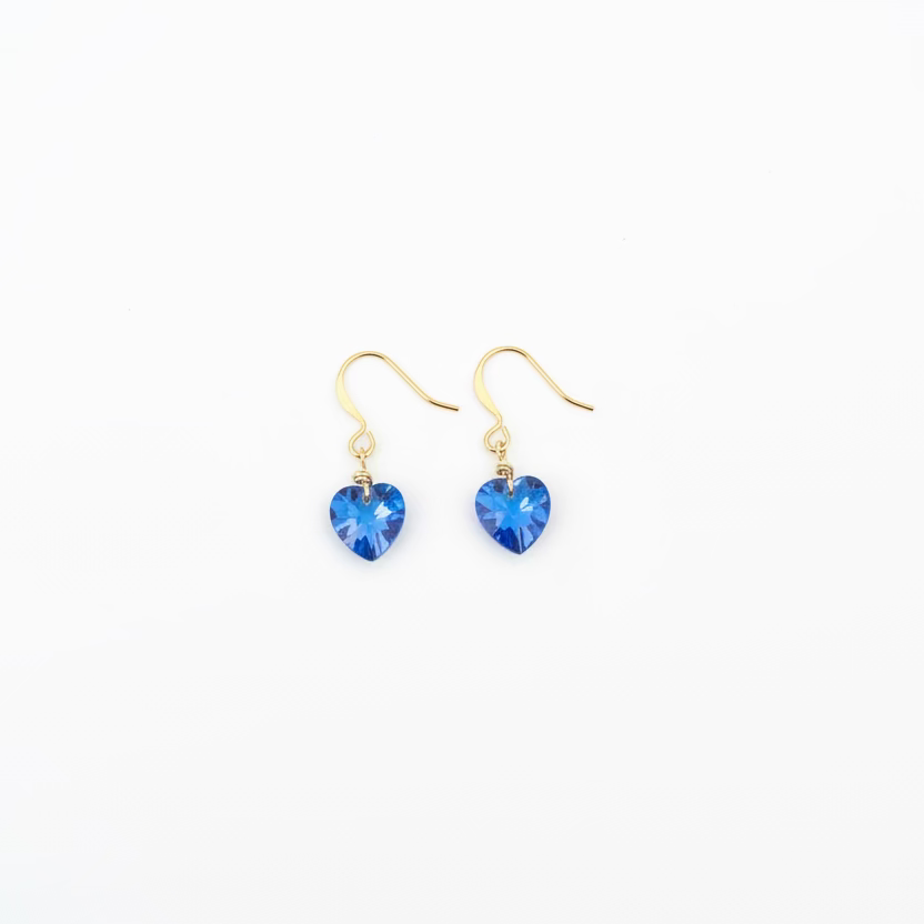 A pair of gold-tone dangle earrings with small, faceted blue heart-shaped crystals, displayed on a light wood grain surface.