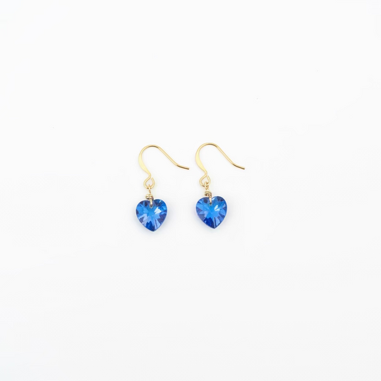 A pair of gold-tone dangle earrings with small, faceted blue heart-shaped crystals, displayed on a light wood grain surface.