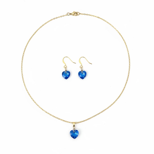 A gold chain necklace and matching drop earrings with blue heart-shaped crystals, displayed on a light wood grain surface.
