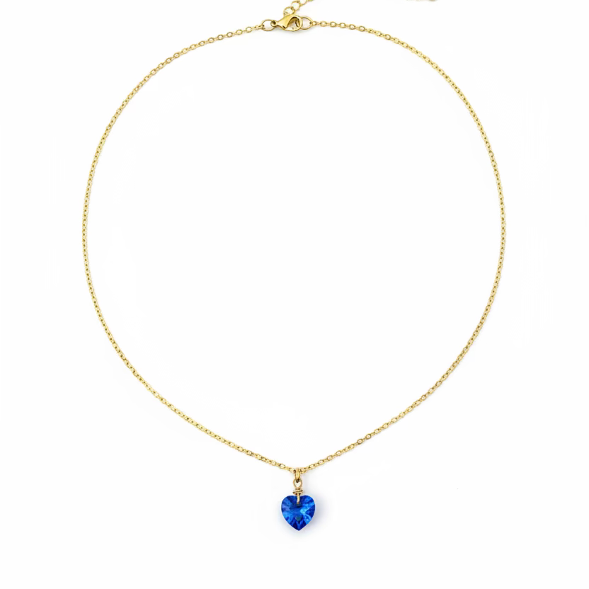 A delicate gold chain necklace with a small blue heart-shaped pendant, displayed on a light wood grain surface.