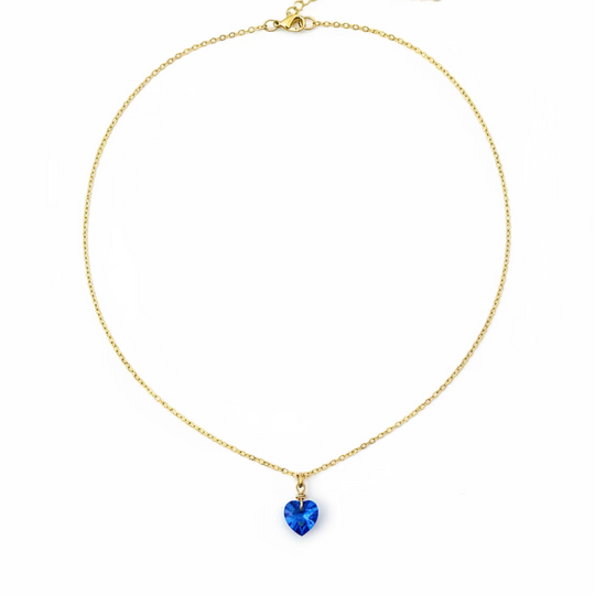 A delicate gold chain necklace with a small blue heart-shaped pendant, displayed on a light wood grain surface.