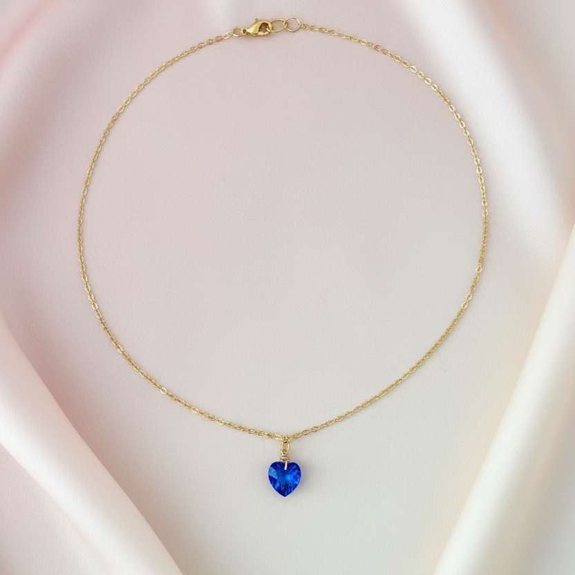 A delicate gold chain necklace with a small, faceted blue heart-shaped pendant, displayed on a light wood grain surface.