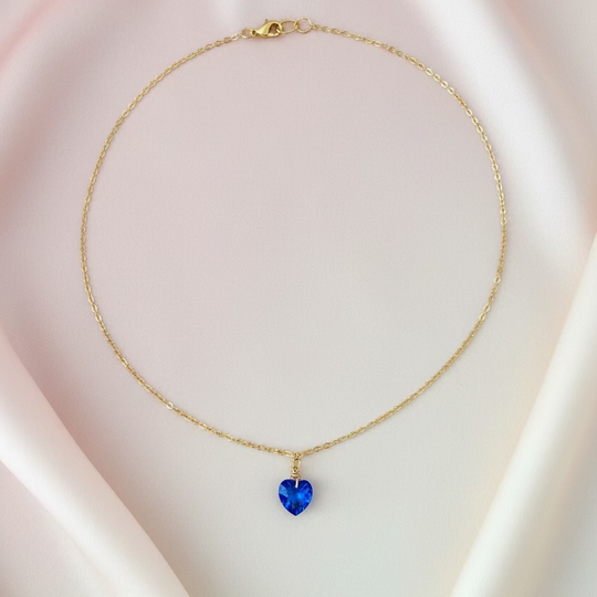 A delicate gold chain necklace with a small, faceted blue heart-shaped pendant, displayed on a light wood grain surface.