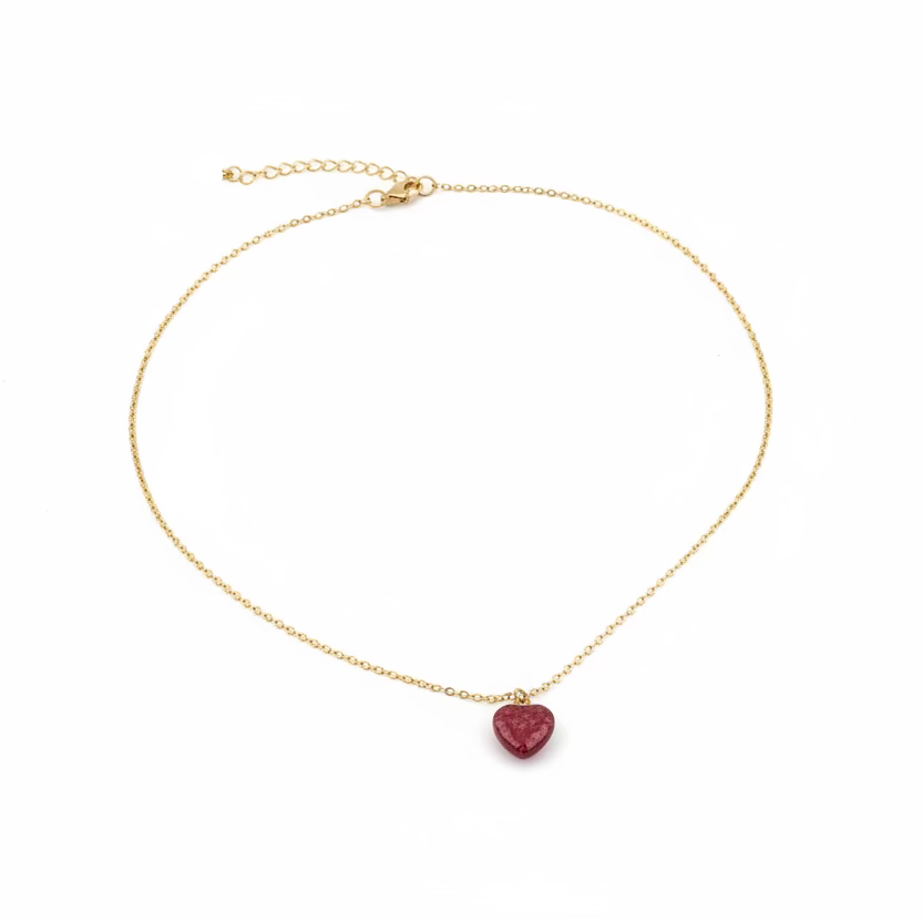 A delicate gold chain necklace with a small heart-shaped red gemstone pendant, displayed on a light wood grain surface.