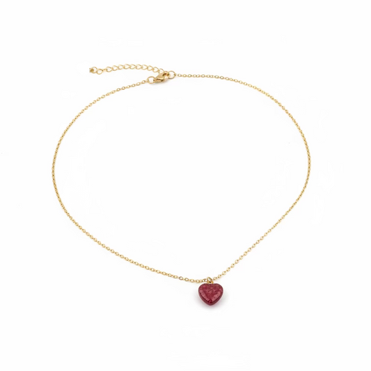 A delicate gold chain necklace with a small heart-shaped red gemstone pendant, displayed on a light wood grain surface.