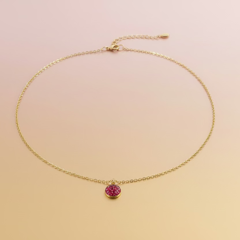 A delicate gold chain necklace with a small, round, dark red gemstone pendant, displayed on a light wood grain surface.