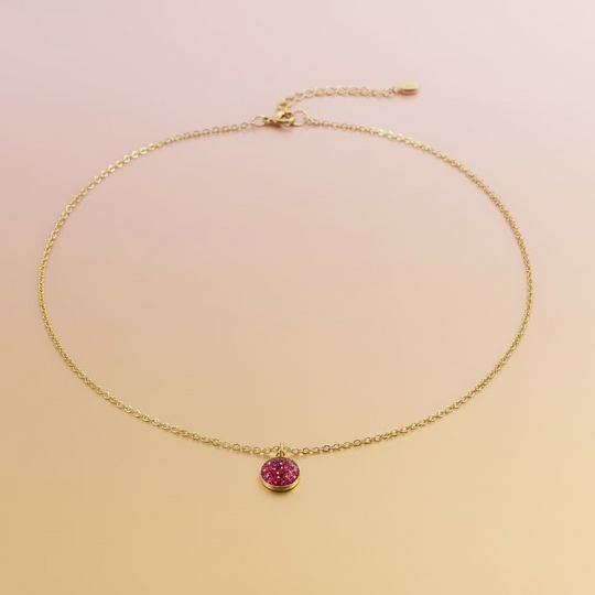 A delicate gold chain necklace with a small, round, dark red gemstone pendant, displayed on a light wood grain surface.