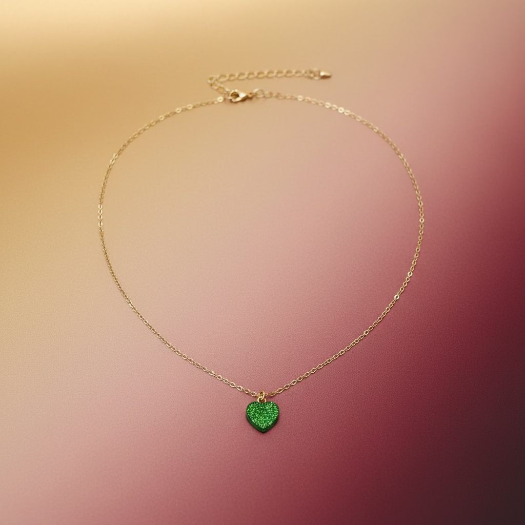 A delicate gold chain necklace with a small, textured green heart-shaped pendant, displayed on a light wood grain surface.