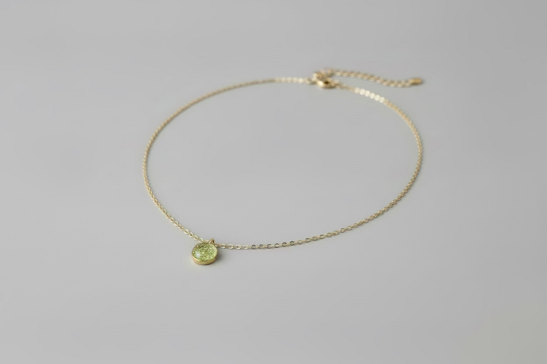 A delicate gold chain necklace with a small, round green pendant, displayed on a light wood grain surface.