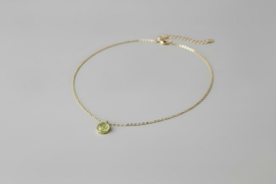 A delicate gold chain necklace with a small, round green pendant, displayed on a light wood grain surface.