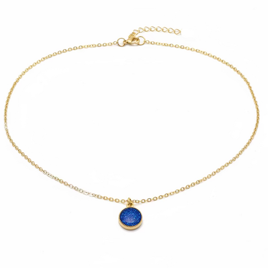 A delicate gold chain necklace with a small, textured circular blue pendant, displayed on a light wood grain surface.