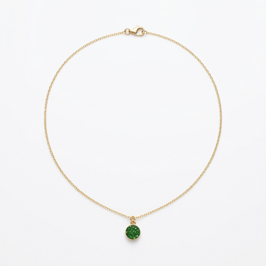 A delicate gold chain necklace with a small round green gemstone pendant, displayed on a textured gray surface with natural light casting shadows.
