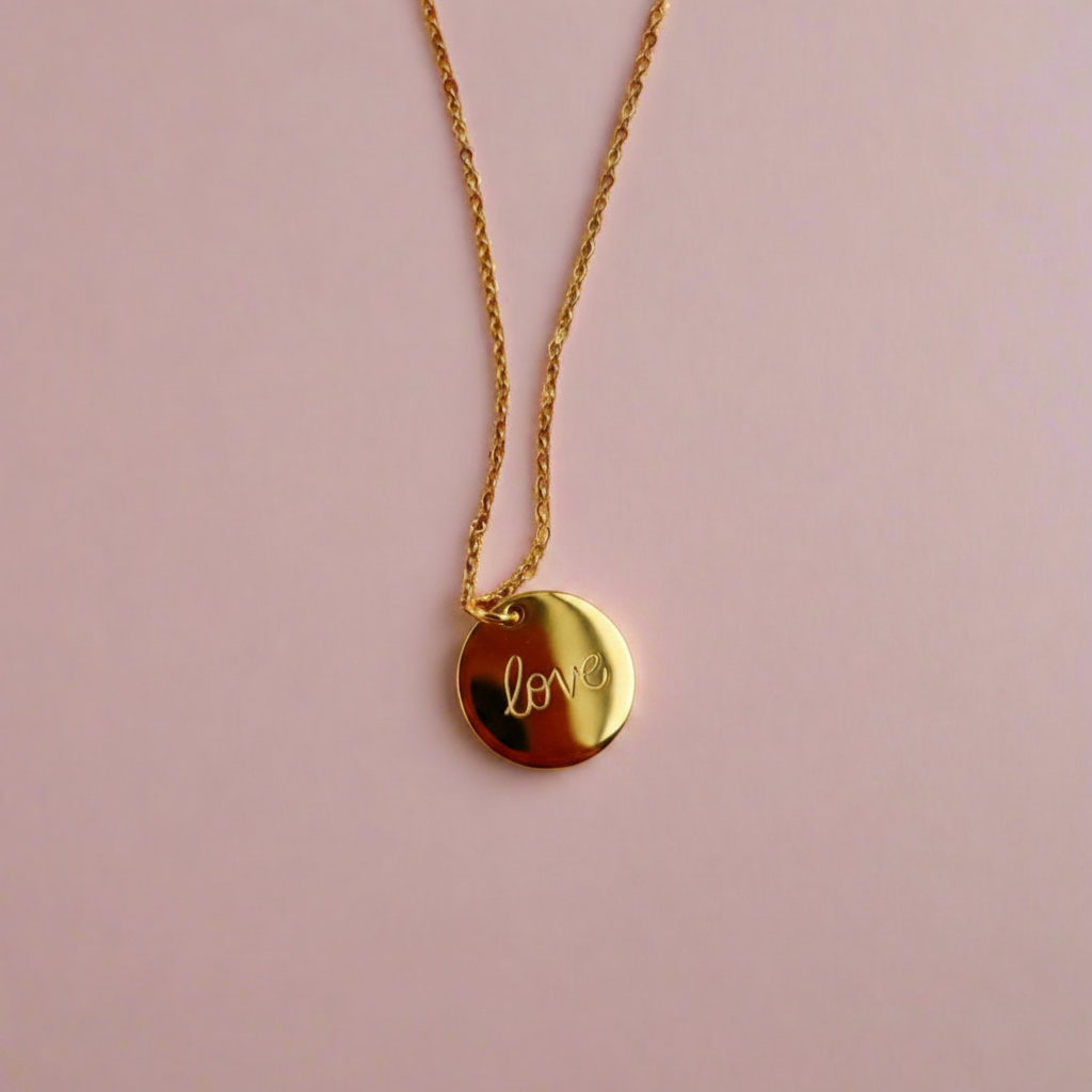 Gold Hand Stamped Love Necklace, our delicate gold disk necklace features a hand stamped "Love" message in simple script. Its minimalist design and 14K gold filled chain add a sweet, subtle touch making it a treasure to adore.
