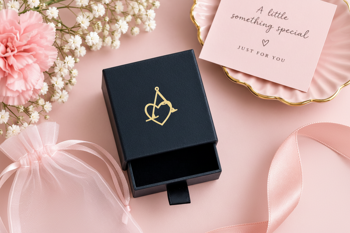 A black jewelry gift box with a gold heart-and-arrow emblem sits open on a pink surface, styled with a cream jewelry pouch, pink carnations, baby's breath, a pink card, and a pink ribbon. The pouch reads 'ALYSSA CHARM JEWELLERY'.